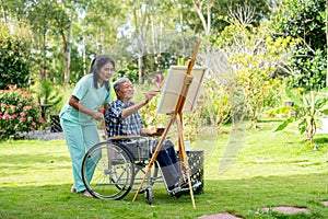 Wide shot of Asian nurse stand beside senior man sit on wheelchair and draw or paint for relax and revive good health of old