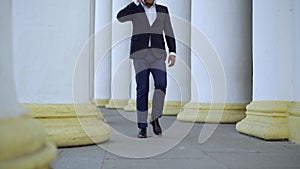 Wide shot of busy African American businessman talking on the phone walking between white columns outdoors and leaving