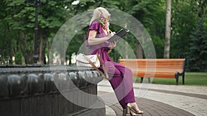 Wide shot busy successful adult Caucasian elegant woman talking on phone writing in sketchbook sitting on fountain in