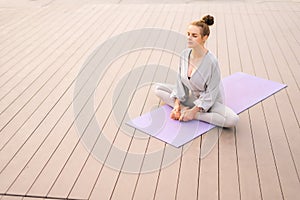 Wide shot of calm young woman sitting on yoga mat with closed eyes and meditating in butterfly pose, tilting head
