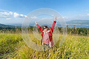 Wide shot of Caucasia woman with red coat and sunglasses spread her arms and stand in the meadow with background of water