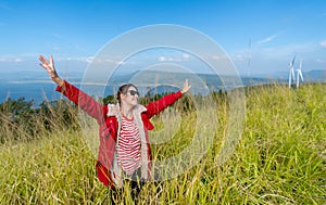 Wide shot of Caucasia woman with red coat and sunglasses spread her arms and stand in the meadow with background of water