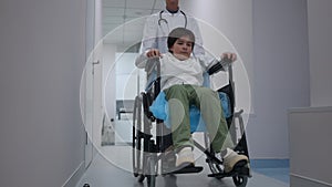 Wide shot Caucasian boy in wheelchair with serious doctor pushing mobility aid device walking in slow motion in hospital