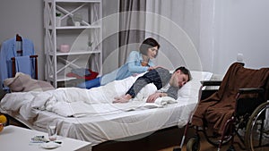 Wide shot Caucasian man lying in bed as loving woman turning body of spouse with paralysis. Portrait of sad husband with