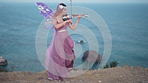 Wide shot Caucasian woman in fairy costume standing on cliff at ocean playing violin. talented confident musician