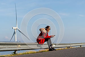 Wide shot of Caucasian woman with red coat and sunglasses sit on barrier of roadside and use mobile phone on mountain near