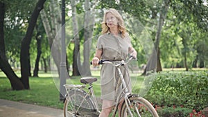 Wide shot of cheerful blond middle aged woman riding bicycle in sunny summer park and stopping. Portrait of excited