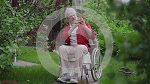 Wide shot of cheerful disabled old man taking selfie with blooming tree in spring summer garden. Portrait of happy