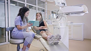 Wide shot cheerful young female dentist and adolescent patient talking using tablet smiling in slow motion. Portrait of