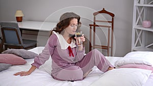 Wide shot confident relaxed lady drinking lemon water smiling looking away sitting on bed at home. Portrait of charming