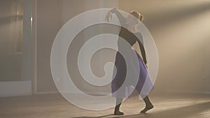 Wide shot of elegant graceful ballerina dancing in backlit fog indoors. Slim beautiful Caucasian young woman performing