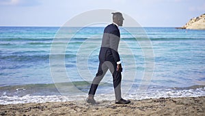 Wide shot of elegant young African American man strolling on sandy beach with turquoise azure sea waves rolling on coast