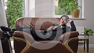 Wide shot of happy Asian groom lying on cozy armchair indoors dreaming on wedding day. Portrait of thoughtful confident