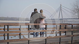 Wide shot of happy Caucasian couple chatting and hugging on riverbank in city park. Back view of loving man and woman