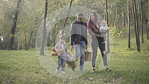 Wide shot of happy Caucasian couple of parents walking with sons on picnic in sunny park. Young beautiful woman and