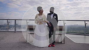 Wide shot of happy newlyweds talking standing at bench on bridge smiling. African American bride and groom holding hands