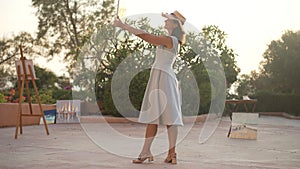Wide shot of happy proud Caucasian female painter dancing with landscape picture in slow motion. Portrait of cheerful