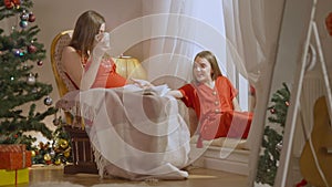 Wide shot of happy relaxed pregnant mother and teenage daughter talking and smiling on Christmas eve at home. Portrait