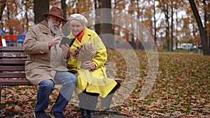 Wide shot happy senior couple sitting on bench on the right talking messaging in smartphone app. Relaxed Caucasian man