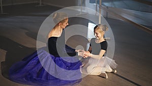 Wide shot of happy slim gorgeous professional ballerina and inspired little girl sitting on floor in ballet studio