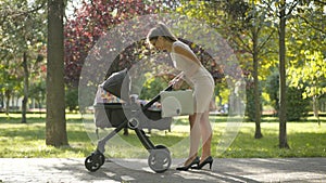 Wide shot of happy young slim woman in elegant dress talking to infant in baby carriage, checking time, and leaving