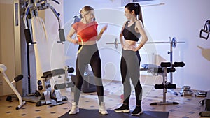 Wide shot of happy young slim woman measuring waist in gym, rejoicing and giving high-five to smiling friend. Portrait