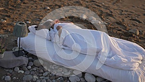 Wide shot of happy young woman sleeping in cushioned white bed on sandy beach. Relaxed carefree Caucasian brunette