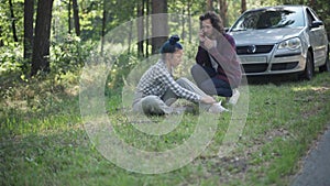 Wide shot injured woman sitting on grass with man running from car calling ambulance. Knocked down Caucasian road