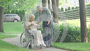 Wide shot of loving old husband giving gift box to paraplegic wife in sunny park. Portrait of affectionate Caucasian man