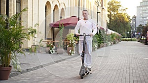 Wide shot millennial man riding electric scooter on city street in slow motion. Portrait of confident handsome young