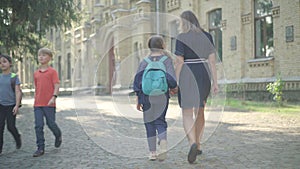 Wide shot of mother and daughter walking along sunny schoolyard and talking. Back view of positive schoolgirl with