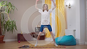 Wide shot of motivated skinny young man jumping doing aerobics exercise at home in 1990s. Positive Caucasian 1980s guy