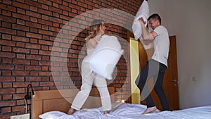 Wide shot pillow fight of joyful Caucasian young couple standing on bed. Cheerful man and woman laughing out loud