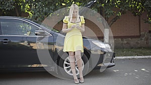 Wide shot portrait of blond stylish Caucasian woman standing at broken car with open hood and messaging on smartphone