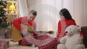 Wide shot portrait of brunette man playing guitar and singing serenade to smiling pregnant woman hugging teddy bear