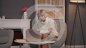 Wide shot portrait of busy Caucasian senior woman surfing Internet on laptop sitting on armchair in living room