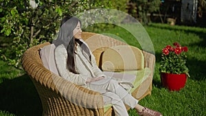 Wide shot portrait of cheerful young gorgeous Asian woman sitting on couch in backyard garden enjoying sunny spring day