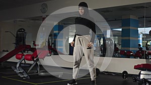 Wide shot portrait of confident slim sportive Middle Eastern woman in hijab doing squats in gym. Motivated athletic