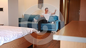 Wide shot portrait of confident young Caucasian man sitting on couch in hotel room with coffee cup looking away thinking