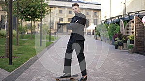 Wide shot portrait of elegant handsome young man in suit standing with skateboard on city street looking at camera