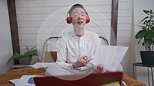 Wide shot portrait of embarrassed Asian man in headphones discussing business strategy with colleagues online from home