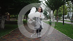 Wide shot portrait of happy confident little person in rain coat standing with balloons outdoors smiling. Carefree