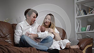 Wide shot portrait of happy couple of parents sitting with newborn baby son on comfortable couch in living room at home