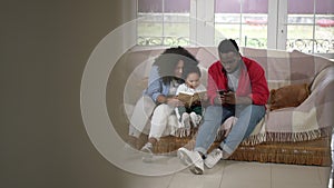 Wide shot portrait loving African American mother reading book to cute son as father messaging online on smartphone