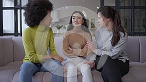 Wide shot portrait of sad brokenhearted beautiful Asian woman sitting on couch as African American and Caucasian women