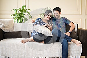 wide shot Portrait shot of happy smiling indian couple busy using mobile phone while sitting on sofa at home - concept