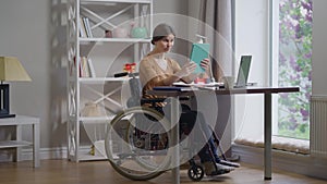 Wide shot portrait smiling beautiful young woman in wheelchair waving talking at tablet video chat sitting at table