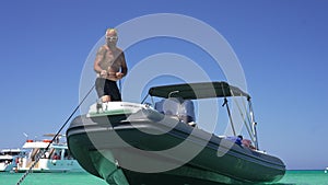 Wide shot portrait of tanned tattooed man pulling anchor chain in slow motion standing on yacht in lagoon. Confident