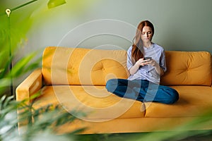 Wide shot portrait of upset young woman using phone, sitting alone on sofa at home, receiving, reading bad news, message