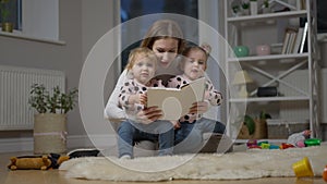 Wide shot portrait of young Caucasian mother reading book for twin daughters sitting in living room. Happy charming